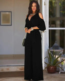 Black cold-shoulder jumpsuit with V-neck and tie waist, worn by a model, full length.