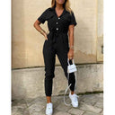 Black short-sleeve jumpsuit with belt and white buttons, front view against a stone wall.