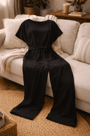Black wide-leg jumpsuit with belted waist and short sleeves, draped on a sofa, angled view.