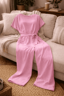 Pink wide-leg belted jumpsuit with tied waist, draped over a light-colored sofa, front view.
