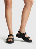 Black chunky platform sandals with three buckle straps worn on feet, angled view showing lug sole and slingback detail.