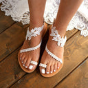 White floral appliqué flat sandals with scalloped lace-trim straps and toe-post construction on tan sole, front view on wooden surface.