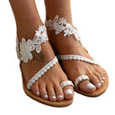 Floral appliqué flat sandals with white lace upper and tan sole, front angled view on neutral background showing full strap and ankle detail.