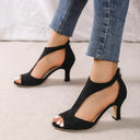 Black suede-touch peep-toe heeled sandals with cutout T-bar upper and back zip closure, front angled view.