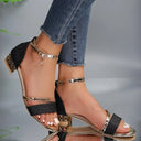 Black glitter block heel sandals with gold metallic ankle strap, crystal charm pendant, and geometric faceted block heel — angled side view on model wearing jeans.