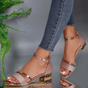 Rose gold glitter block heel sandals with metallic ankle strap and crystal charm pendant, worn with jeans — angled front lifestyle view.
