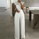 White jumpsuit with lace yoke and pleated wide legs, full front view in a modern room.