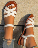 White criss-cross strap flat sandals with tan sole and contrast stitching, overhead view on textured ground surface.