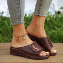 Brown cushioned slip-on low wedge sandal with wide perforated strap and gold-tone buckle accent, angled front view on wooden surface.