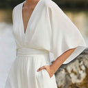 White dolman sleeve jumpsuit with V-neckline and belted waist, hand in pocket, angled view.
