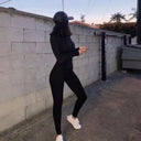 Black slim-fit jumpsuit with long sleeves, shown from the side against a brick wall.