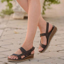 Black cushioned wedge sandals with cork-effect wedge sole and adjustable ankle strap, side view on paved outdoor surface.