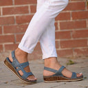 Blue cushioned wedge sandals with adjustable ankle strap worn with white trousers, side view against brick wall background.
