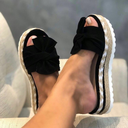 Black platform wedge sandals with fabric bow detail, woven braided midsole and cream stitched rubber outsole, angled side view.