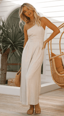 Beige one-shoulder jumpsuit with cinched waist, full-body view on a patio.