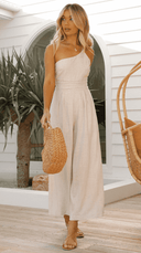 Beige one-shoulder jumpsuit with cinched waist, full front view on a wooden deck.
