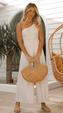 Beige one-shoulder jumpsuit with cinched waist, holding a straw bag, front view.