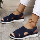 Navy blue mesh low wedge sandals with white cushioned sole and velcro slingback strap, styled on feet with white trousers, angled view.