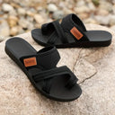 Black arch support flat sandals with mesh cross-strap, angled view on concrete surface showing cushioned footbed.