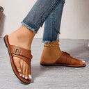Brown tan leather flat toe-ring sandal with buckle cross-strap and contrast stitching, worn with distressed blue jeans, angled front view.