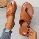Tan leather flat toe-ring sandal with cross-strap and contrast stitching, overhead angled view on foot with gold anklet.
