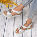 White open-toe espadrille wedge sandals with braided rope sole and elasticated ring-hardware ankle strap, overhead view on whitewashed wood background.